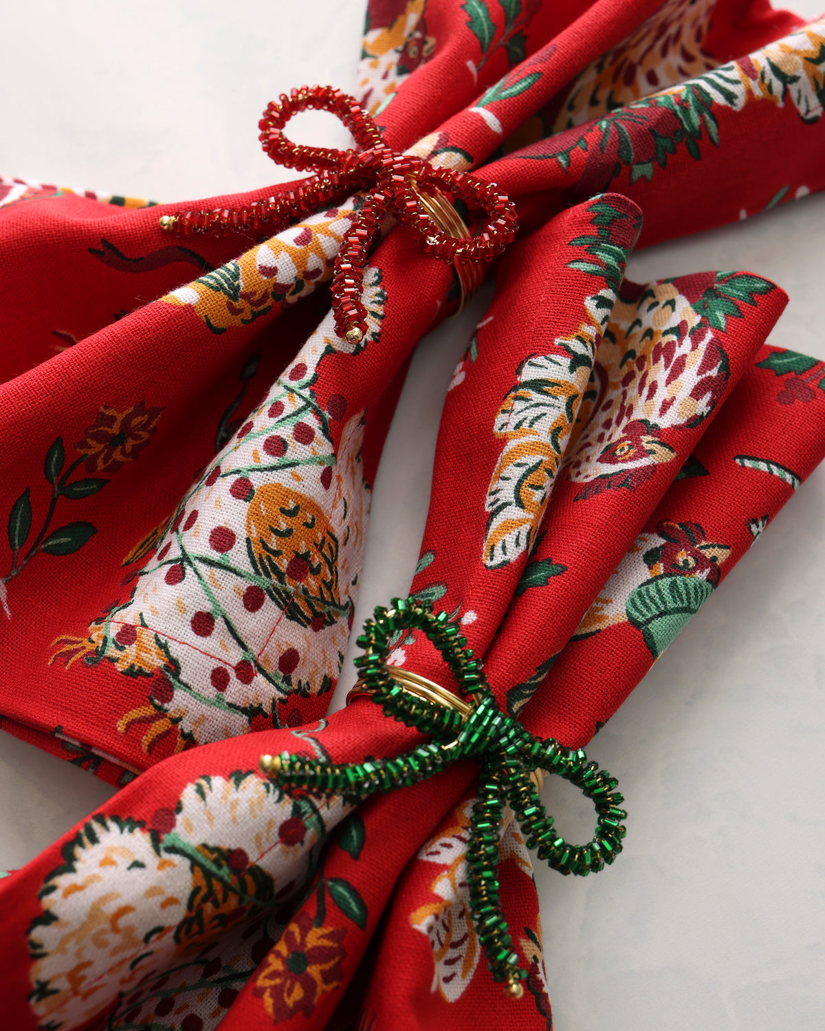 Two beaded bow napkin rings in red and green shown on patterned napkins.