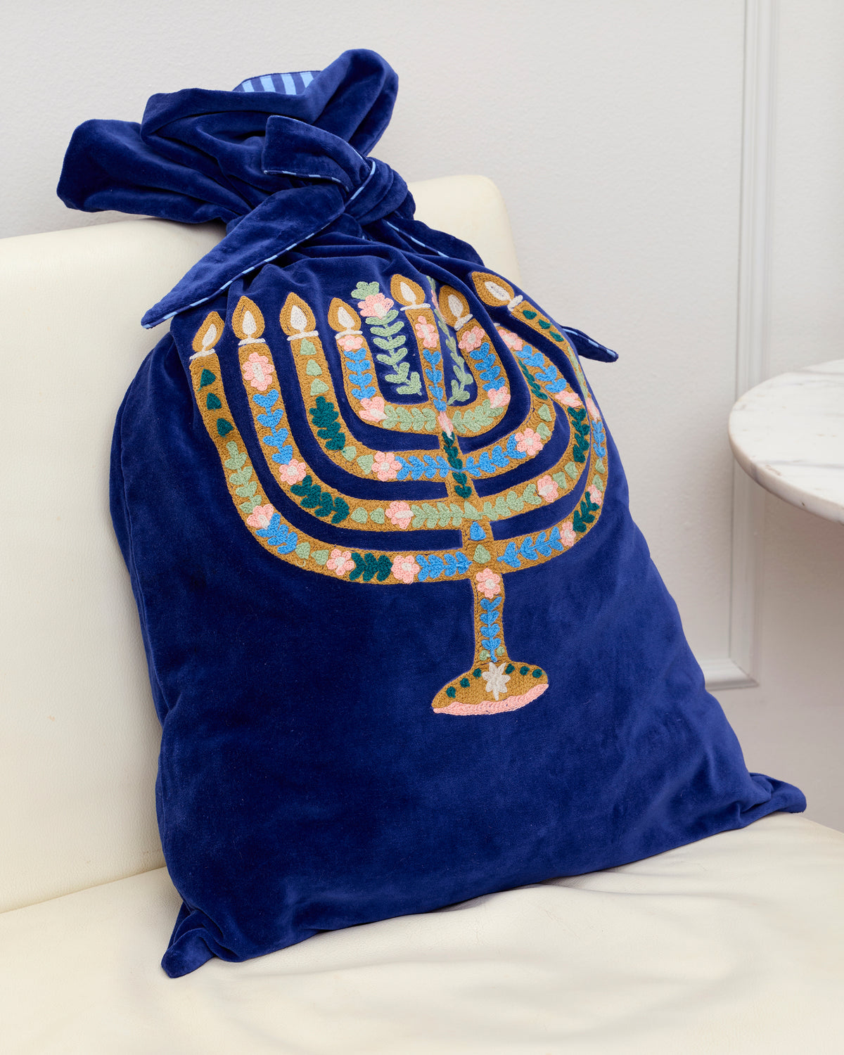 Navy blue embroidered gift sack with a colorful menorah design and coordinating tie, shown on a chair.