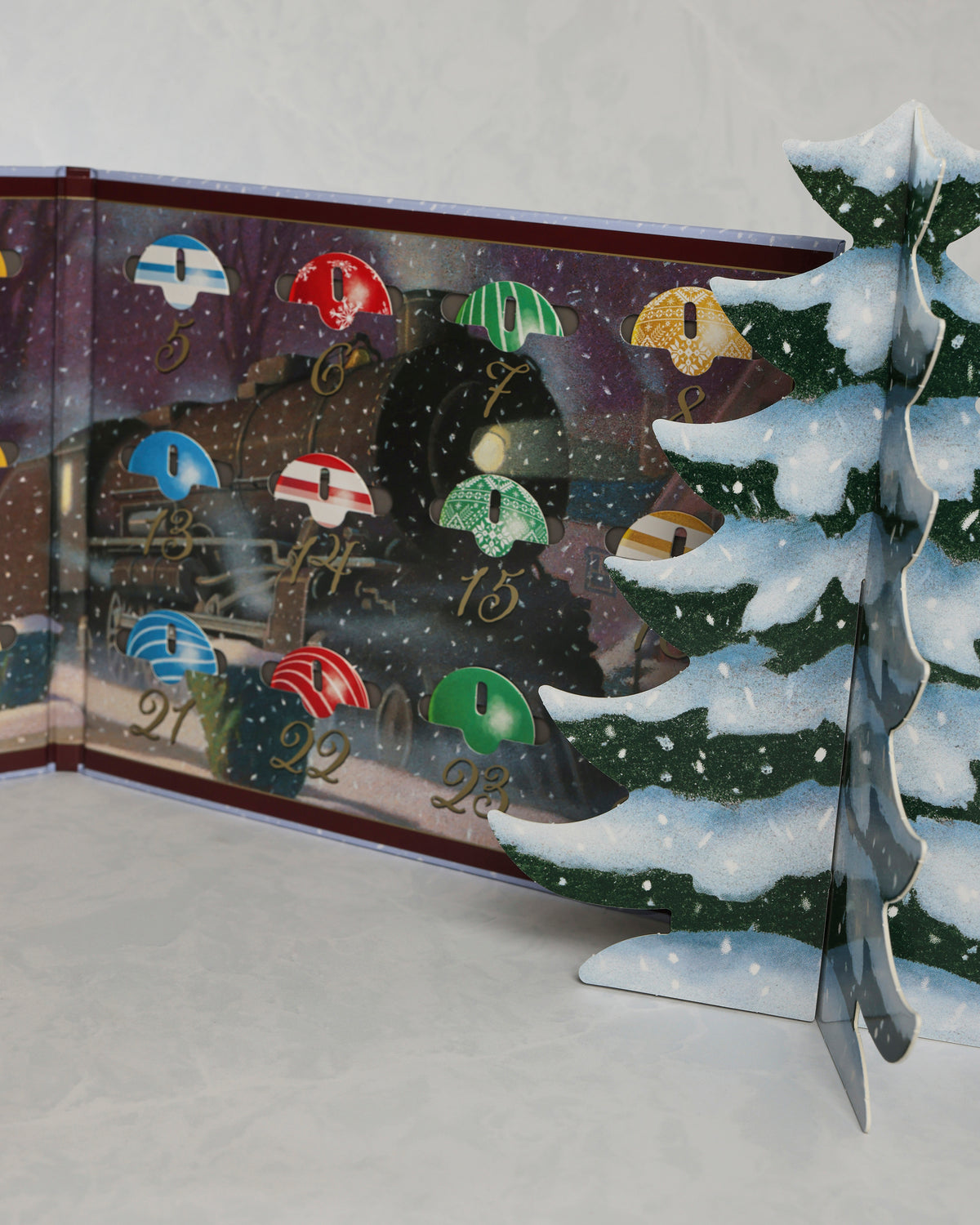 Open Christmas countdown calendar showing numbered ornaments and a freestanding snowy Christmas tree