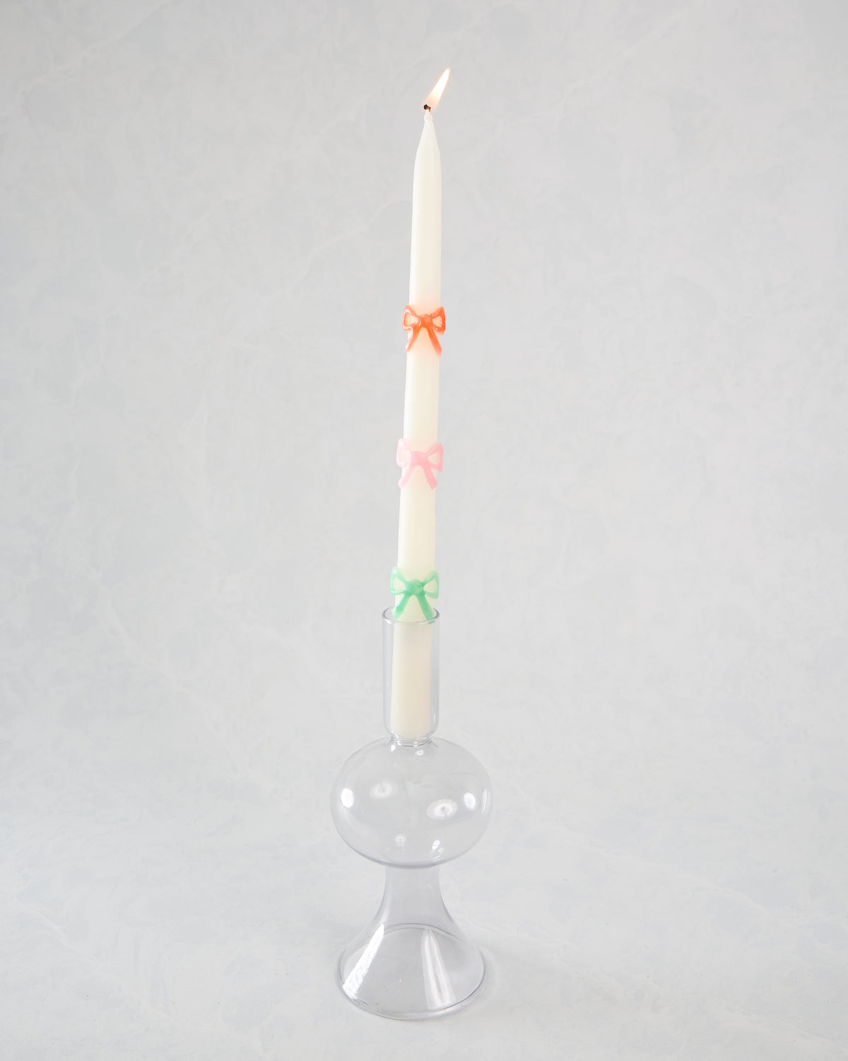 Lit white taper candle with orange, pink, and green bow decorations displayed in a clear glass holder.