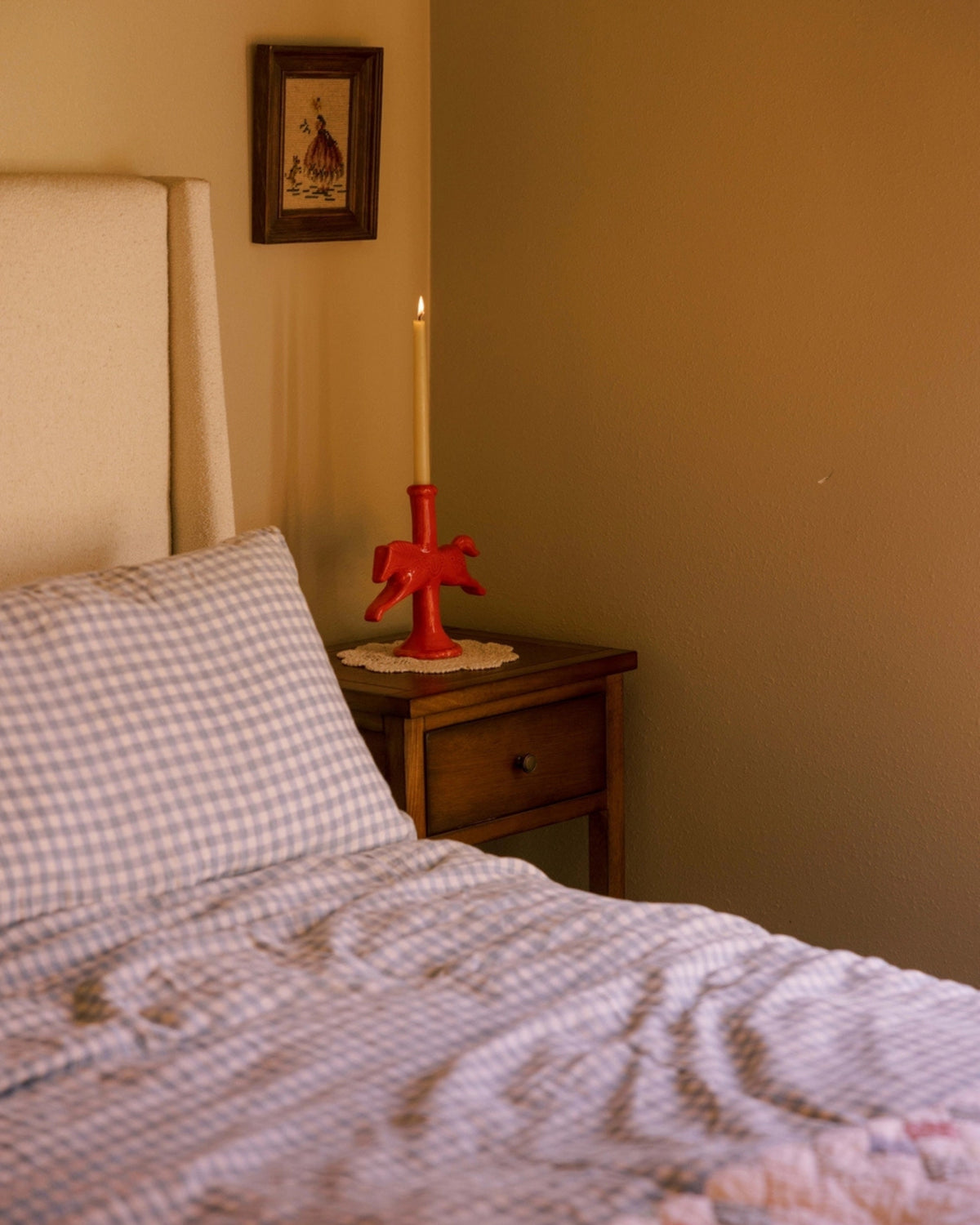 Red horse-shaped candlestick holder with a lit taper candle on a bedside table beside a bed.