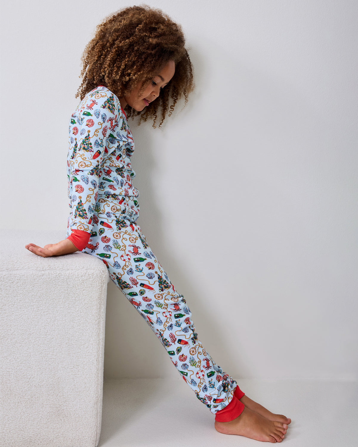 Child wearing a light blue long-sleeve pajama set with a nautical Christmas print, seated beside a white cube.