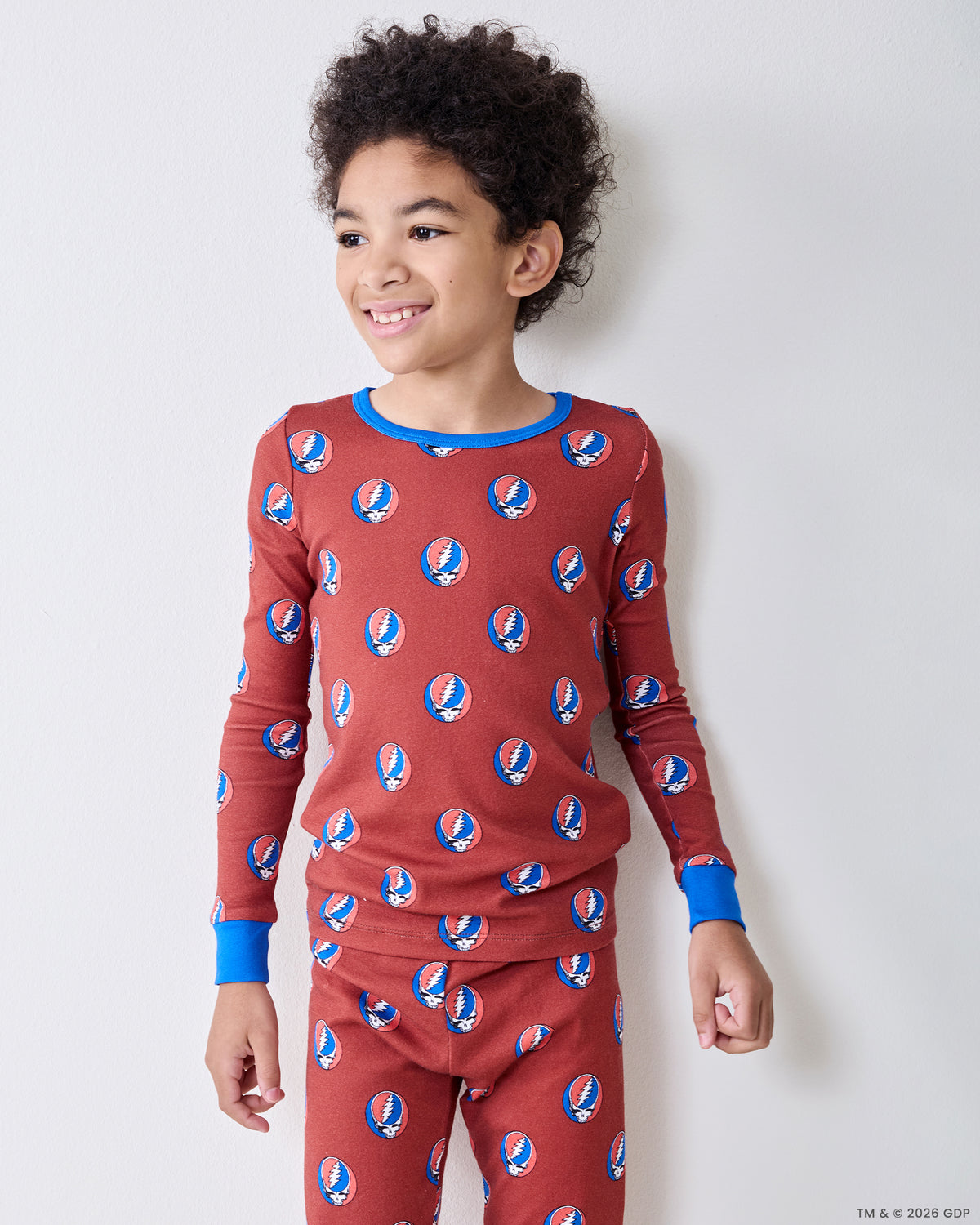 Three-quarter view of a child wearing a long-sleeve red pajama top and matching pants with repeated circular skull-and-lightning-bolt motifs.