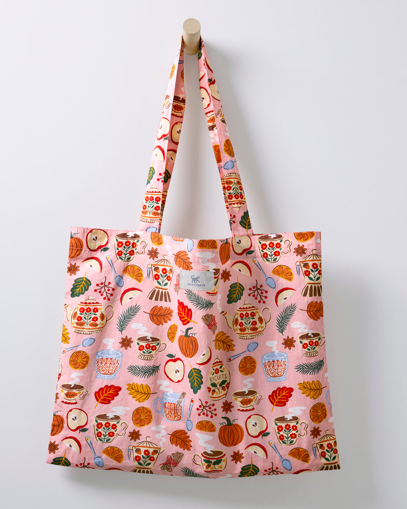 Rose pink tea-themed tote bag hanging from a wooden peg, with long fabric handles.