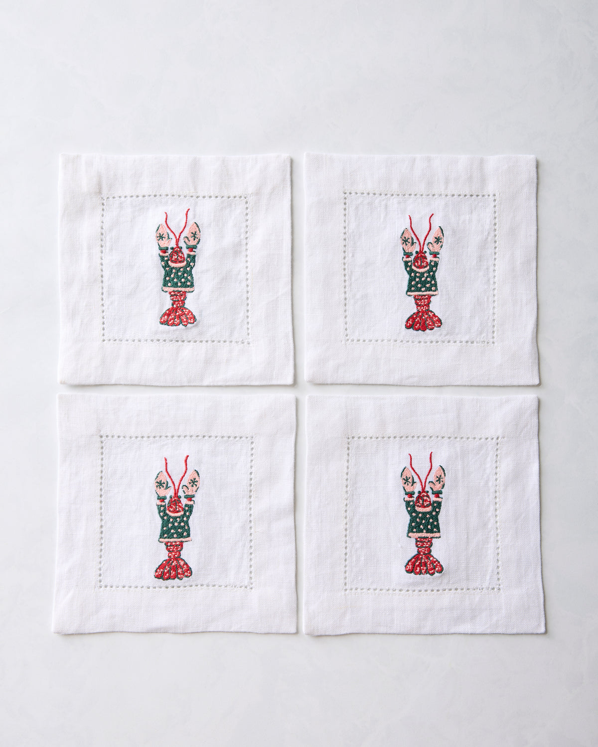 Four white cocktail napkins arranged in a square, each embroidered with a lobster motif.