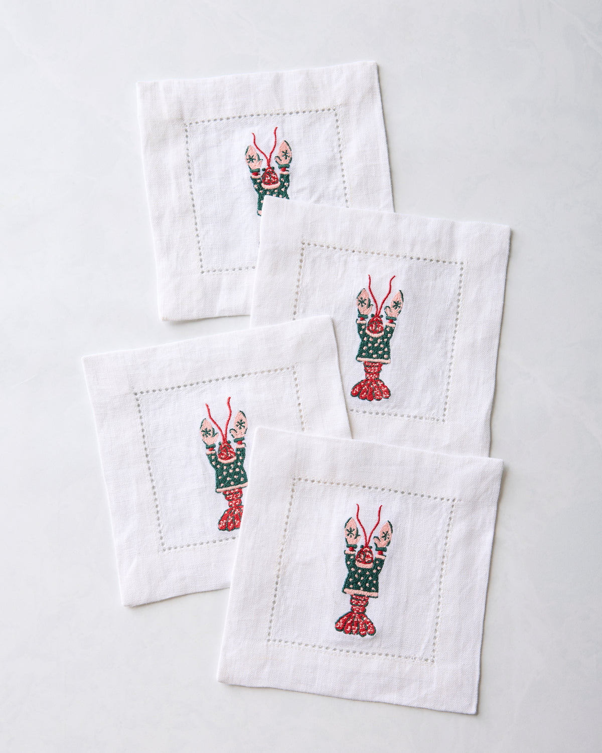 Four white cocktail napkins overlapping in a diagonal arrangement, each with an embroidered lobster motif.
