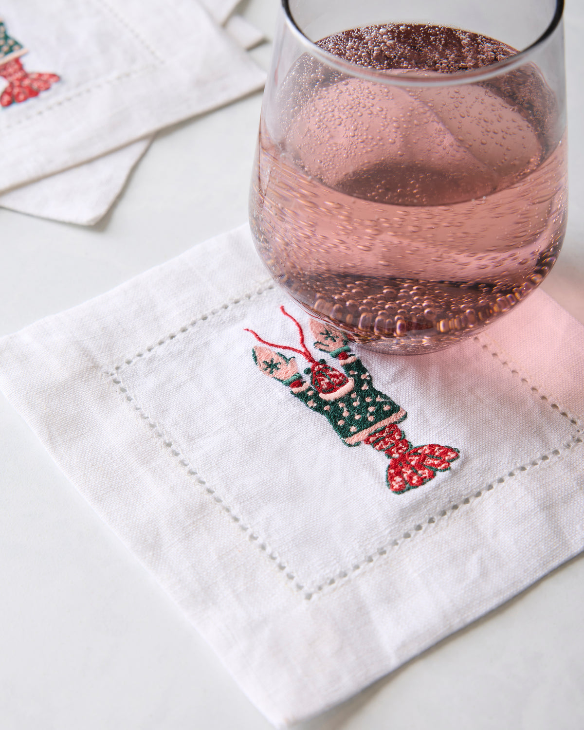 White embroidered cocktail napkin beneath a glass of pink sparkling drink, with additional napkins in the background.