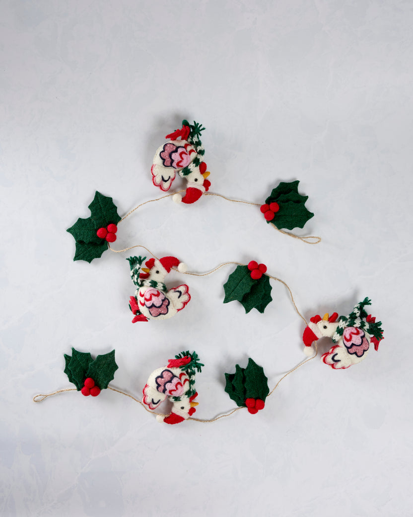 Holiday hens garland loosely coiled on a white surface, showing four felt hens, holly leaves, and red berries.