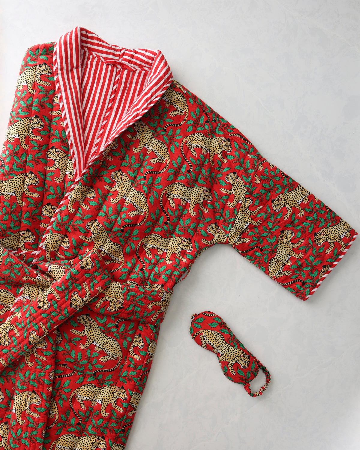 Red quilted robe with a leopard-and-holly print, showing its red-and-white striped lining, alongside a matching eye mask.