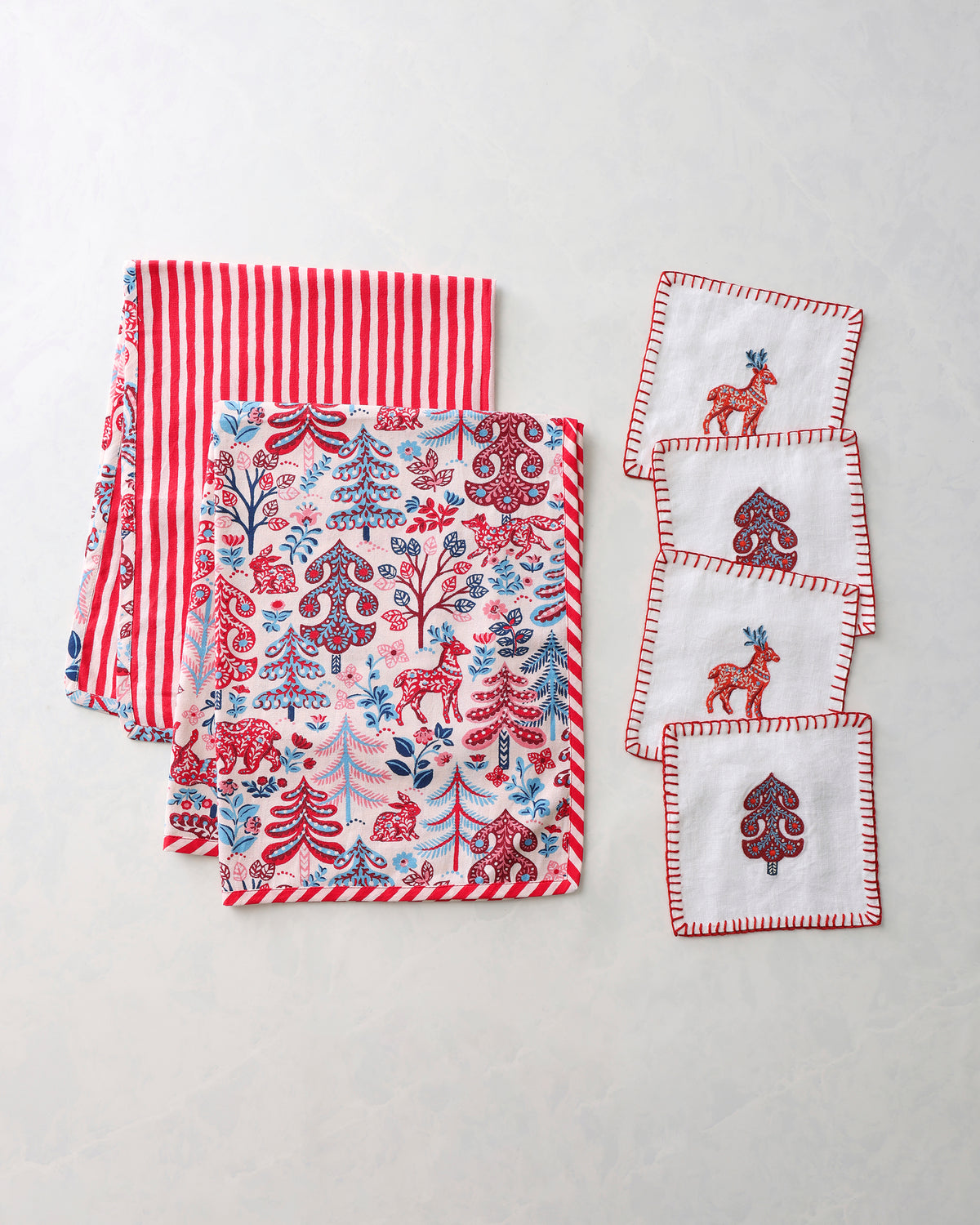 Tea towels and cocktail napkins arranged together, with red-and-white stripes, a forest print, and embroidered motifs.