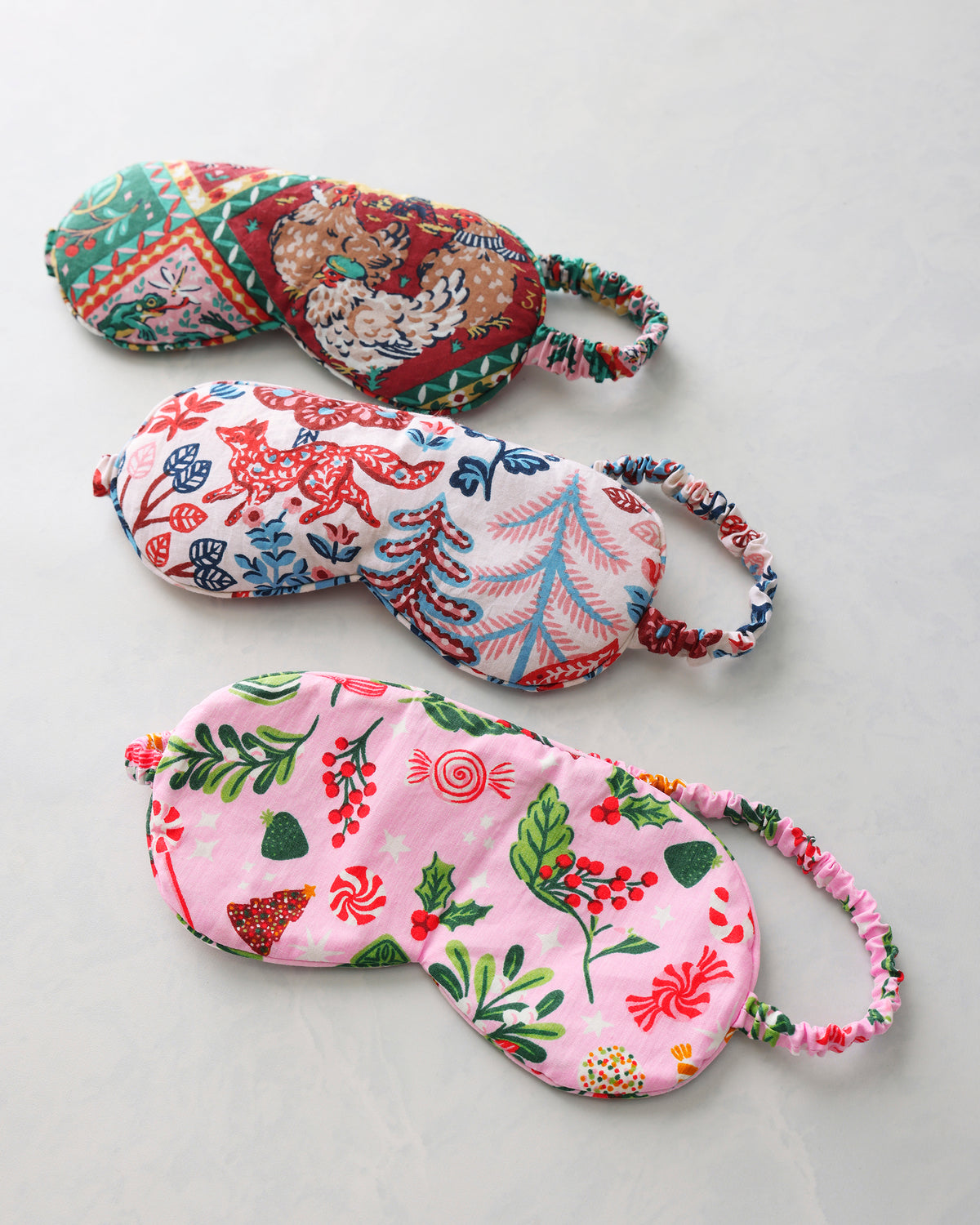 Three printed sleep masks arranged diagonally, featuring a red-and-green holiday pattern, a multicolored woodland pattern, and a pink candy-and-holly pattern.