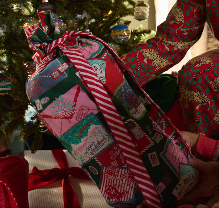 Close-up of a colorful holiday patchwork textile held in front of a decorated Christmas tree; red printed pajamas are visible at right.
