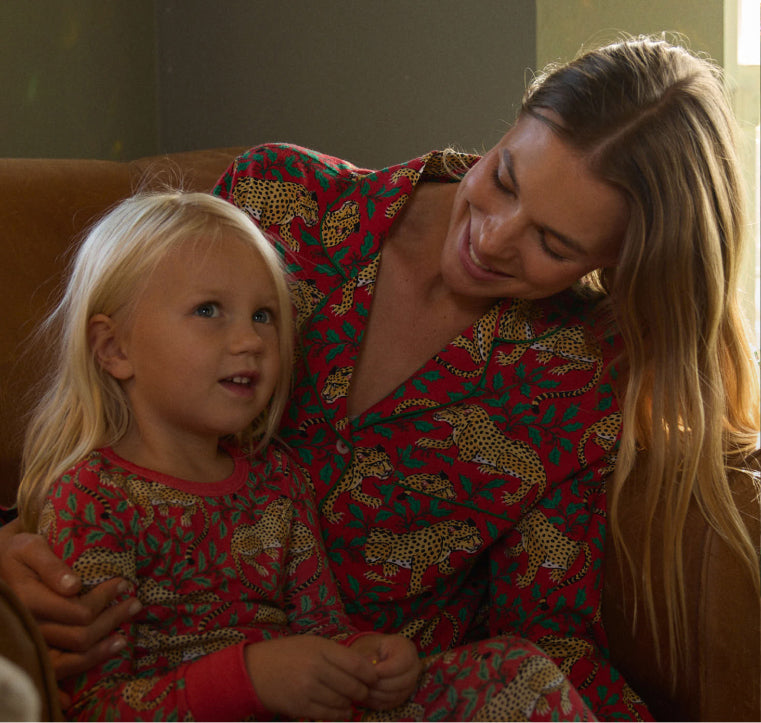 Two people seated together indoors, wearing matching red pajamas with repeating animal and leafy prints.