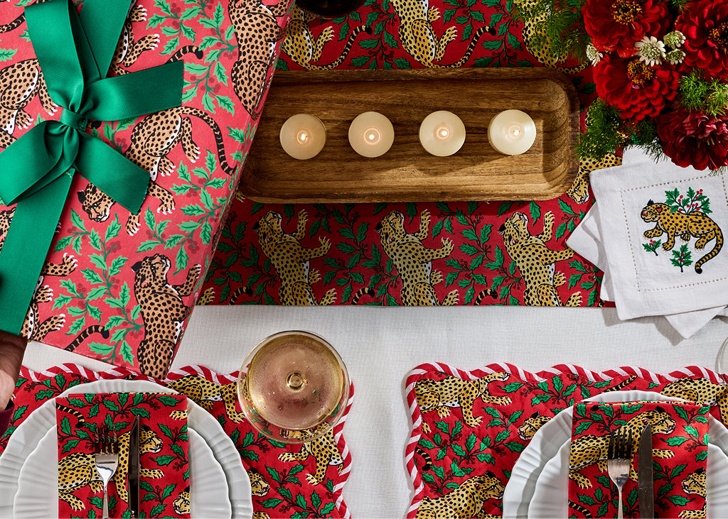 Overhead holiday table setting with red patterned textiles, four candles on a wooden tray, flowers, plates, and wrapped gifts.
