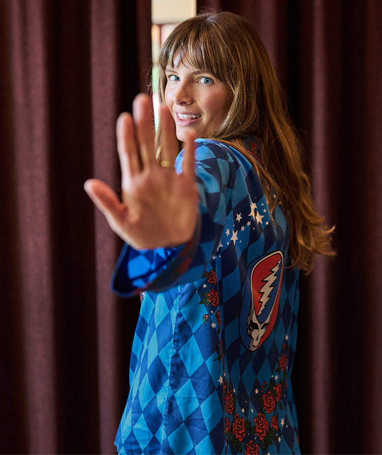 Person in a blue diamond-patterned outfit looks back toward the camera with one hand raised in the foreground; a winged emblem is visible on the back.