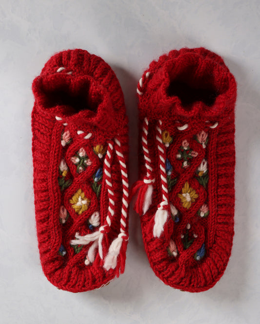 Top view of a pair of red handknit slippers with embroidered floral details and white-and-red ties.