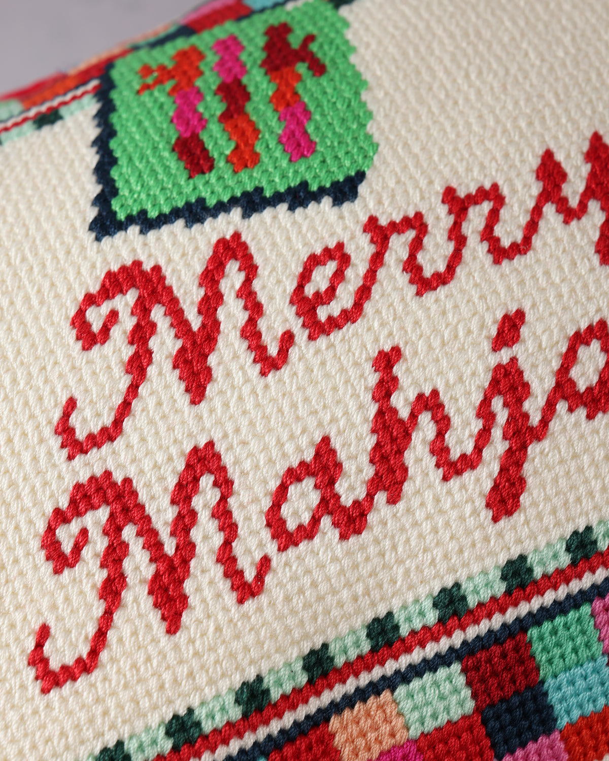 Close-up of the needlepoint pillow showing red “Merry Mahjong” lettering, a green ornament motif, and multicolor border stitching.
