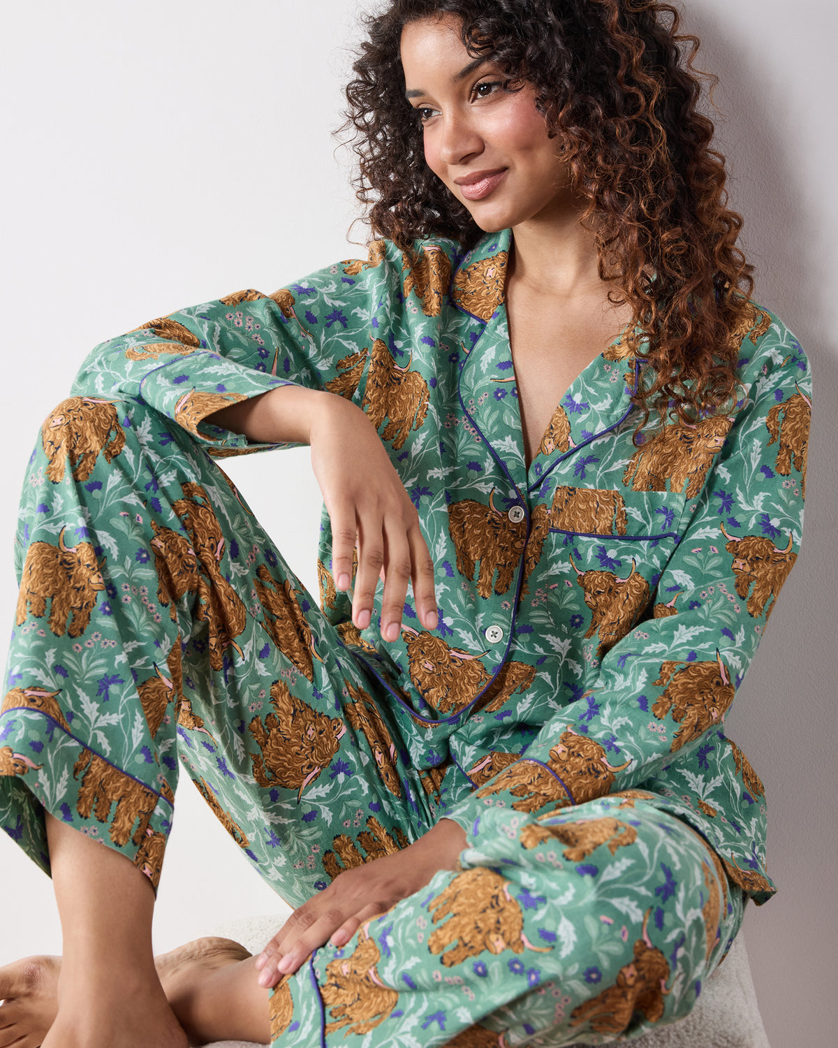 Seated person wearing the Soft Thistle Highland Cows long pajama set, showing the button-down top and matching pants.