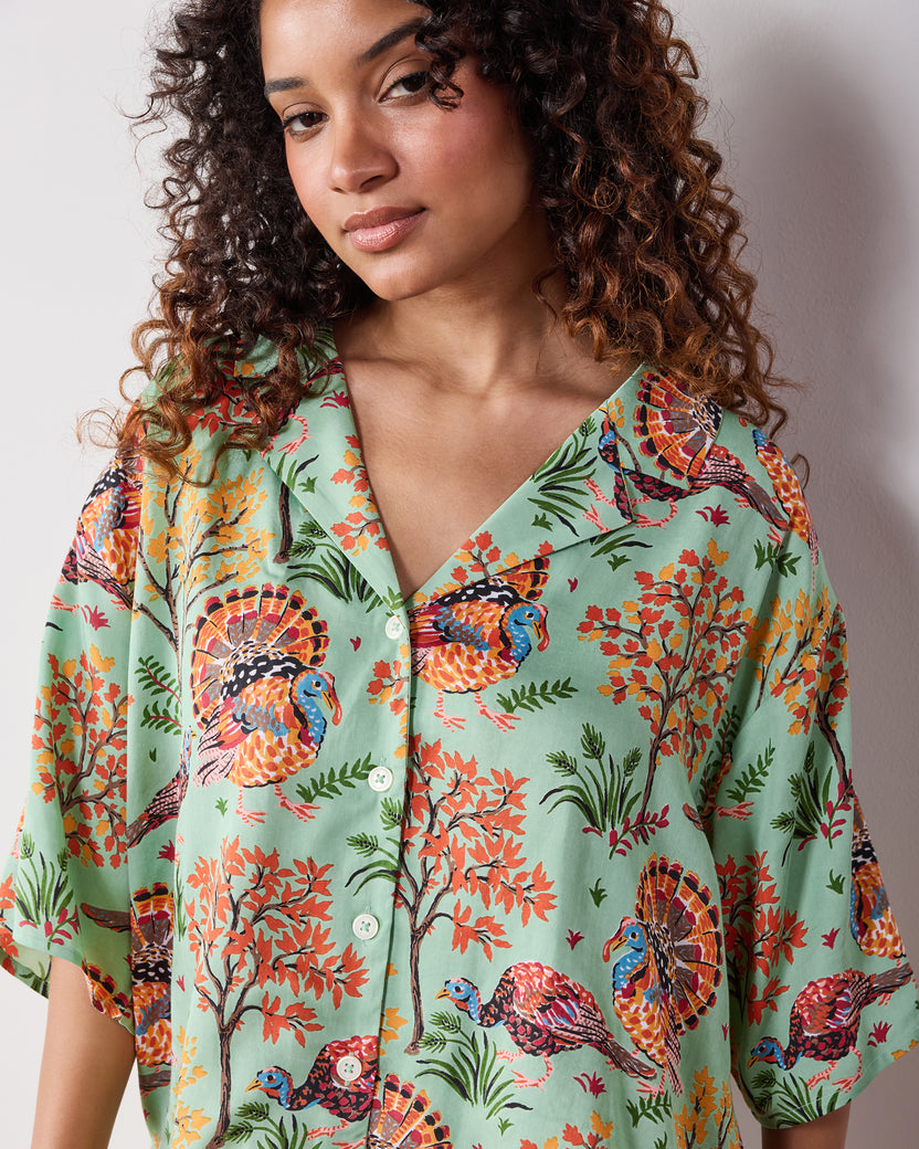 Model wearing a soft sage pajama top printed with colorful turkeys, trees, and foliage.