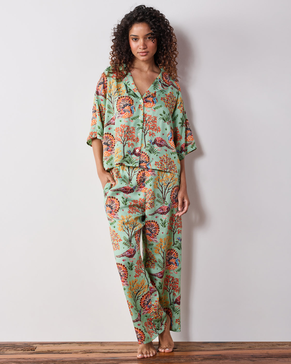 Model wearing a soft sage pajama set with a short-sleeved printed top and wide-leg pants featuring turkeys and foliage.