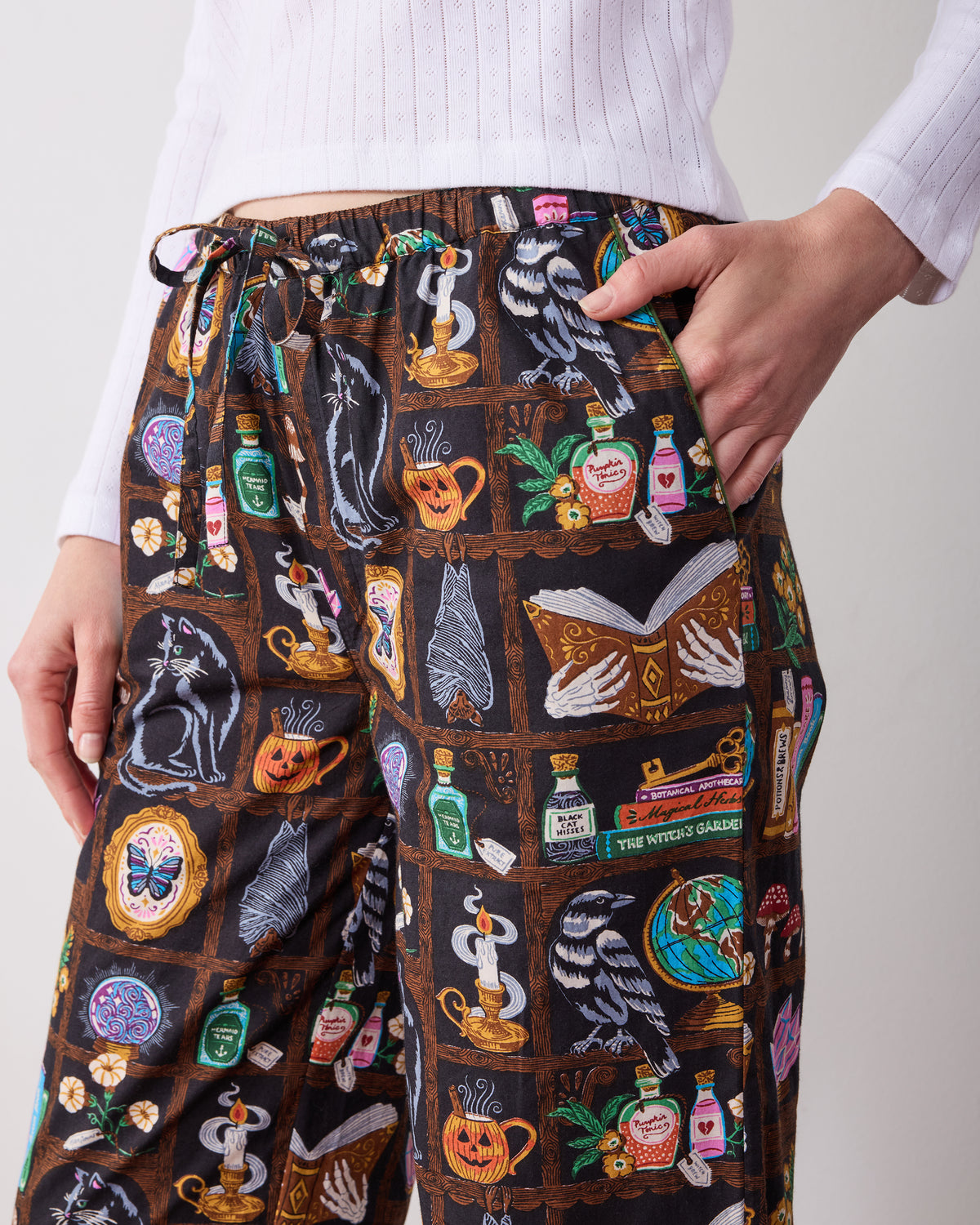 Close front view of the Enchanted Study pajama pants showing the drawstring waistband, side pocket, and illustrated cabinet-of-curiosities print.