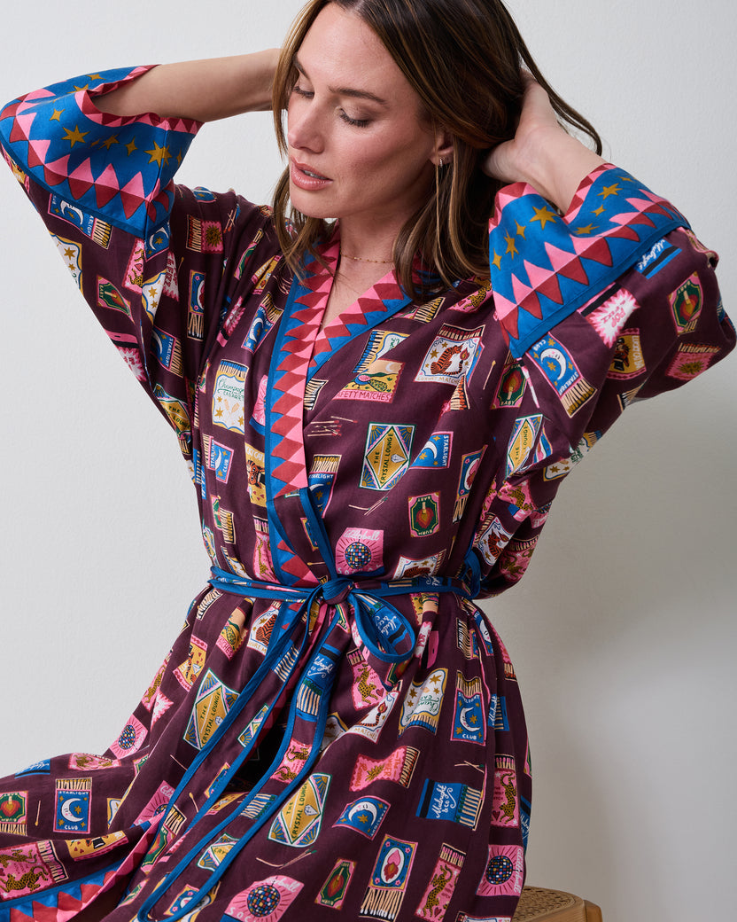 Seated front view of the Cabernet Painted Matchbook robe, showing the wrap front, tied belt, patterned fabric, and contrasting geometric trim on the collar and cuffs.