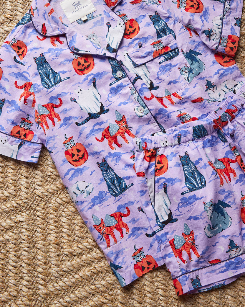 Flat lay close-up of the lavender Halloween House Cats pajama set, showing the piped collar, buttons, chest pocket, elastic waistband, and printed cats, pumpkins, ghosts, and bats.