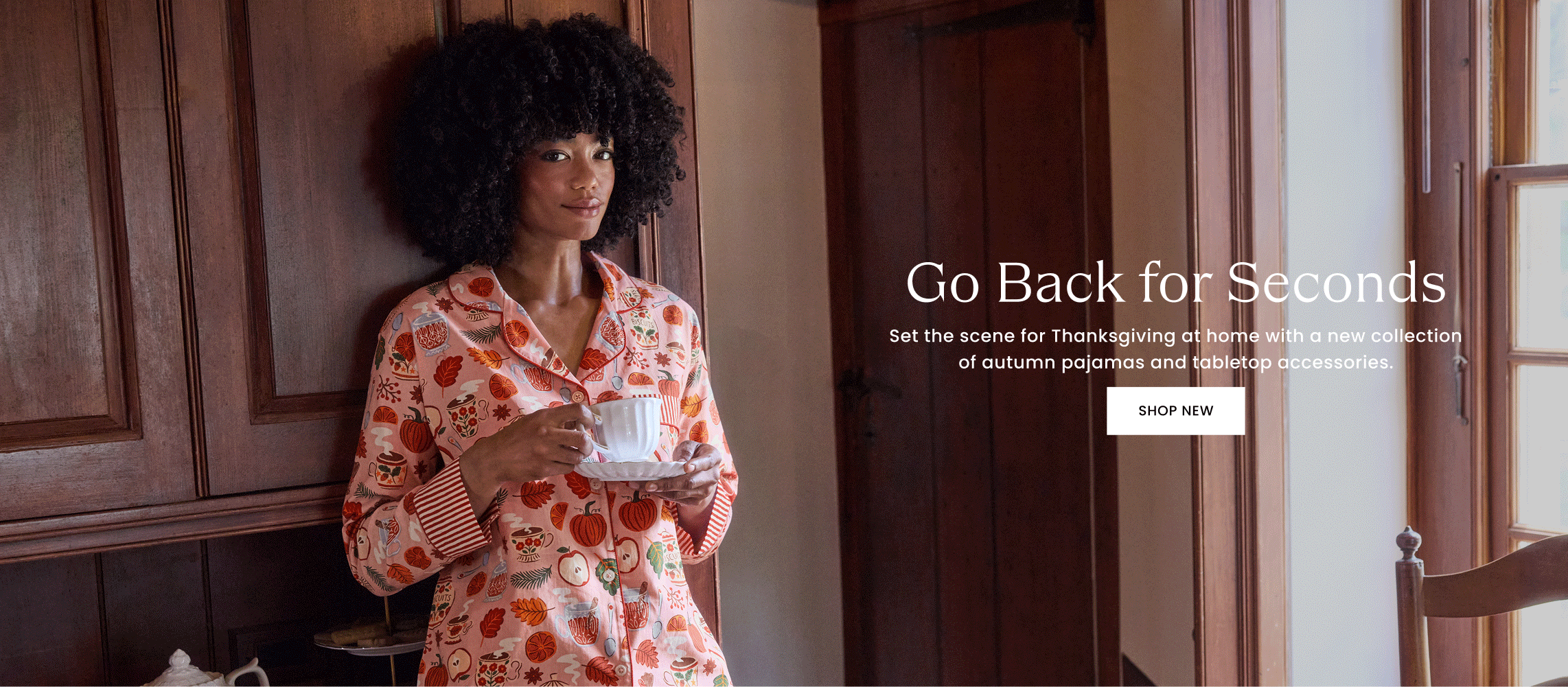 Person in patterned pajamas holding a cup and saucer in a wood-paneled room; banner reads “Go Back for Seconds,” “Set the scene for Thanksgiving at home with a new collection of autumn pajamas and tabletop accessories,” and “SHOP NEW.”