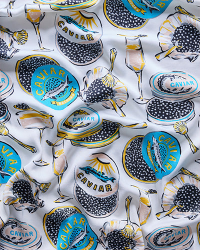 Close-up of the Champagne and Caviar pajama print showing caviar tins, champagne glasses, spoons, and shells on a pale background.