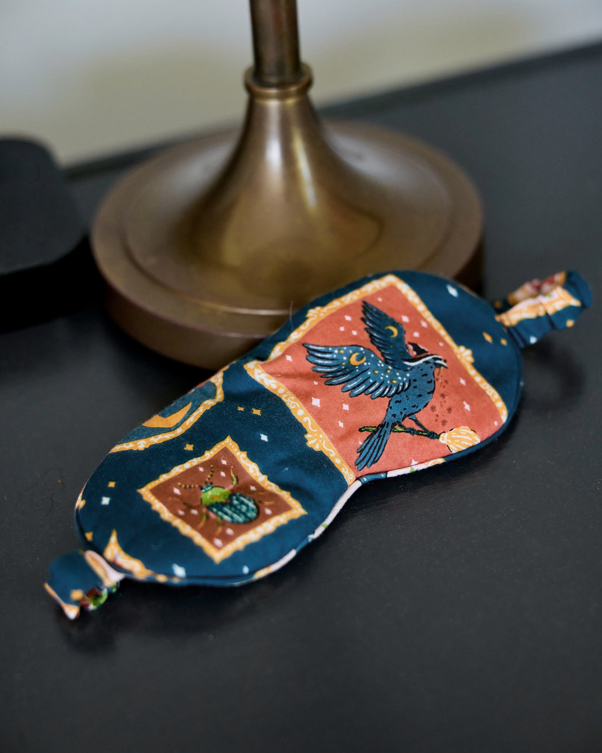 Midnight Gallery eye mask with an adjustable strap, featuring a dark blue print with framed bird and beetle illustrations, shown on a tabletop.