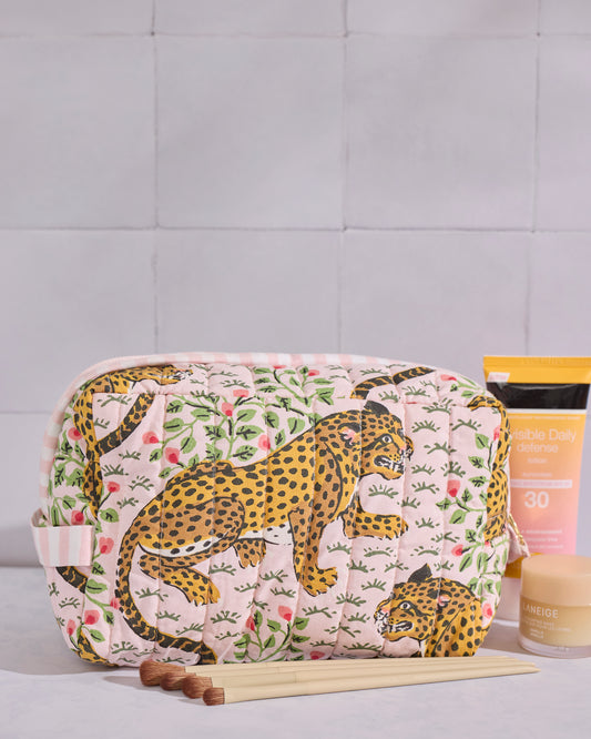 Pink quilted Bagheera pouch with leopard and botanical print, shown beside makeup brushes and skincare products.