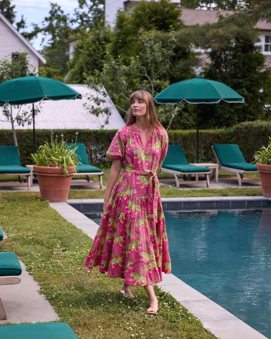 Full-length view of the hot pink Bagheera To and From Dress with leopard print, puff sleeves, tied waist belt, and full tiered skirt beside a swimming pool.