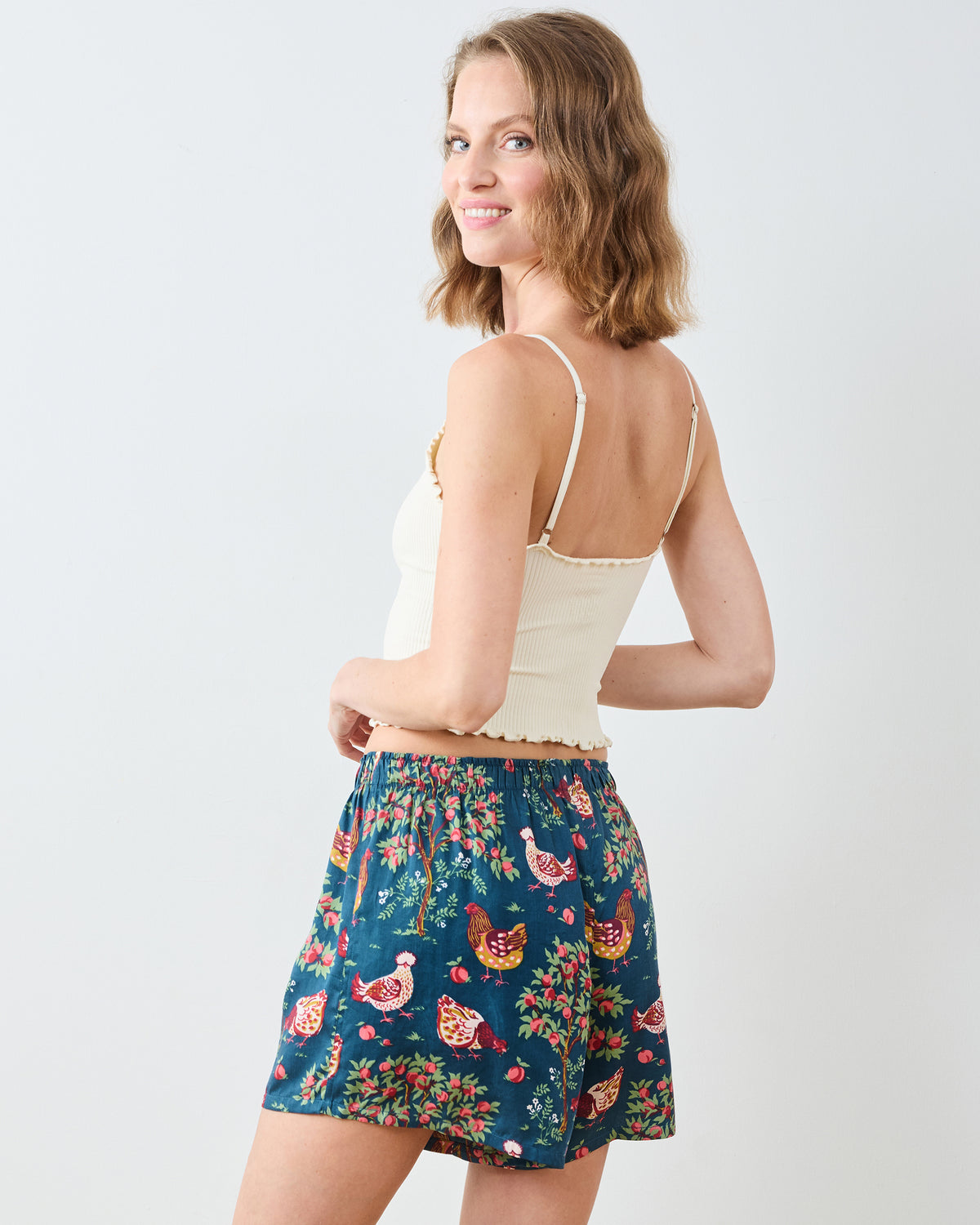 Rear three-quarter view of navy boyfriend boxer shorts with a colorful chicken and garden print, worn with a cream camisole.