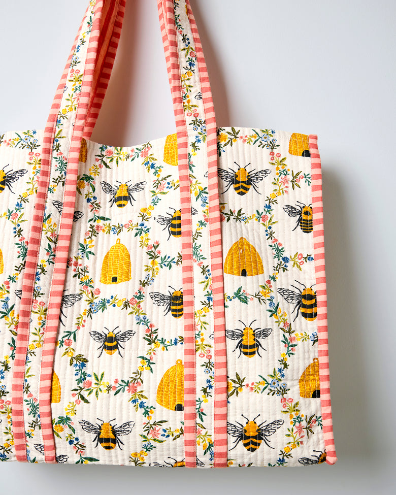 Close view of the cream quilted traveler tote showing bee, beehive, and floral print with pink striped handles and edge trim.