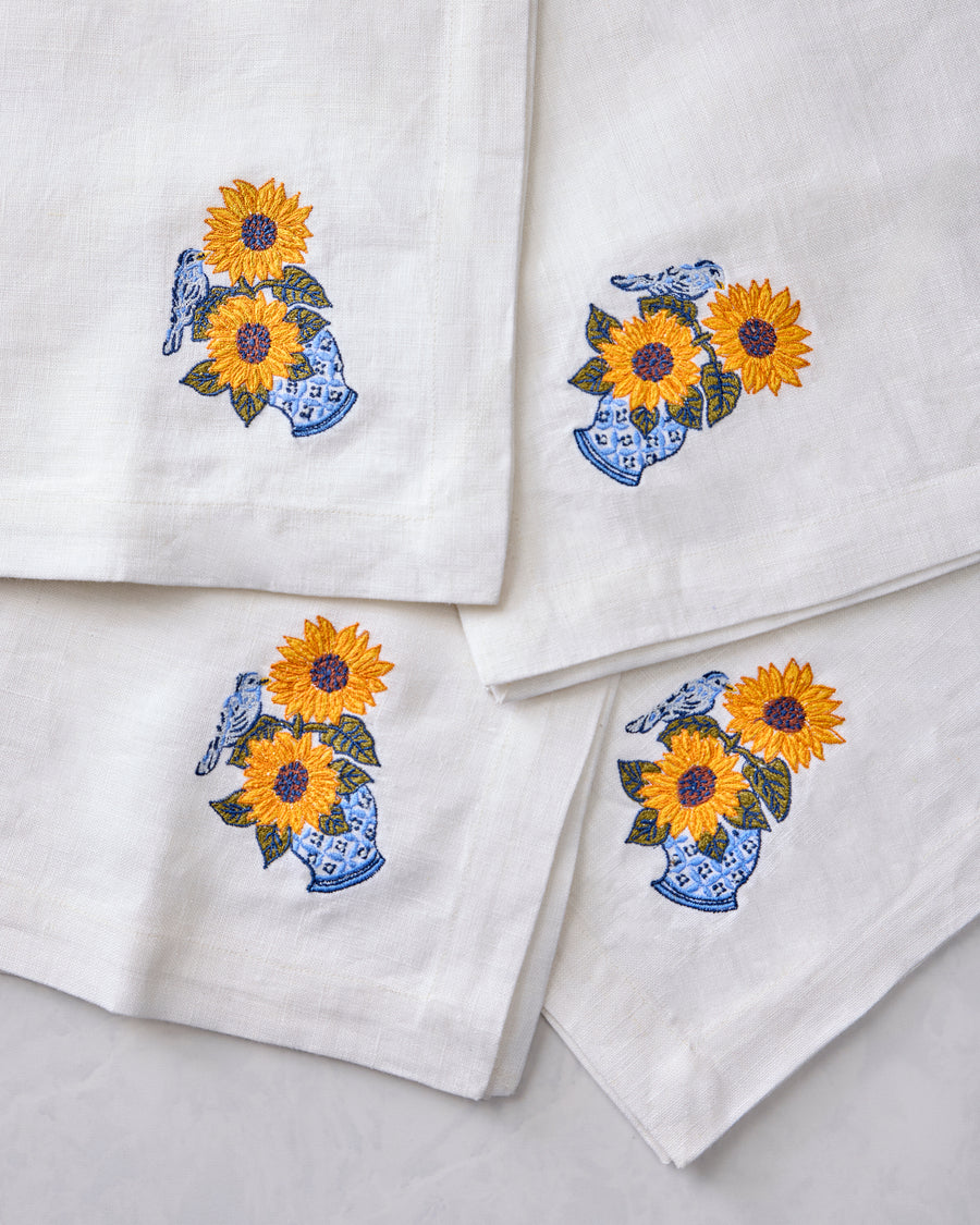 Close-up of four white napkins featuring embroidered sunflowers in blue vases with birds.