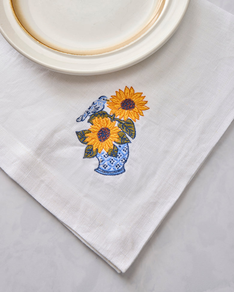 White napkin with embroidered sunflowers in a blue vase and a bird, shown beside a plate.