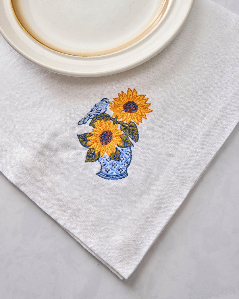 White napkin with embroidered sunflowers in a blue vase and a bird, shown beside a plate.