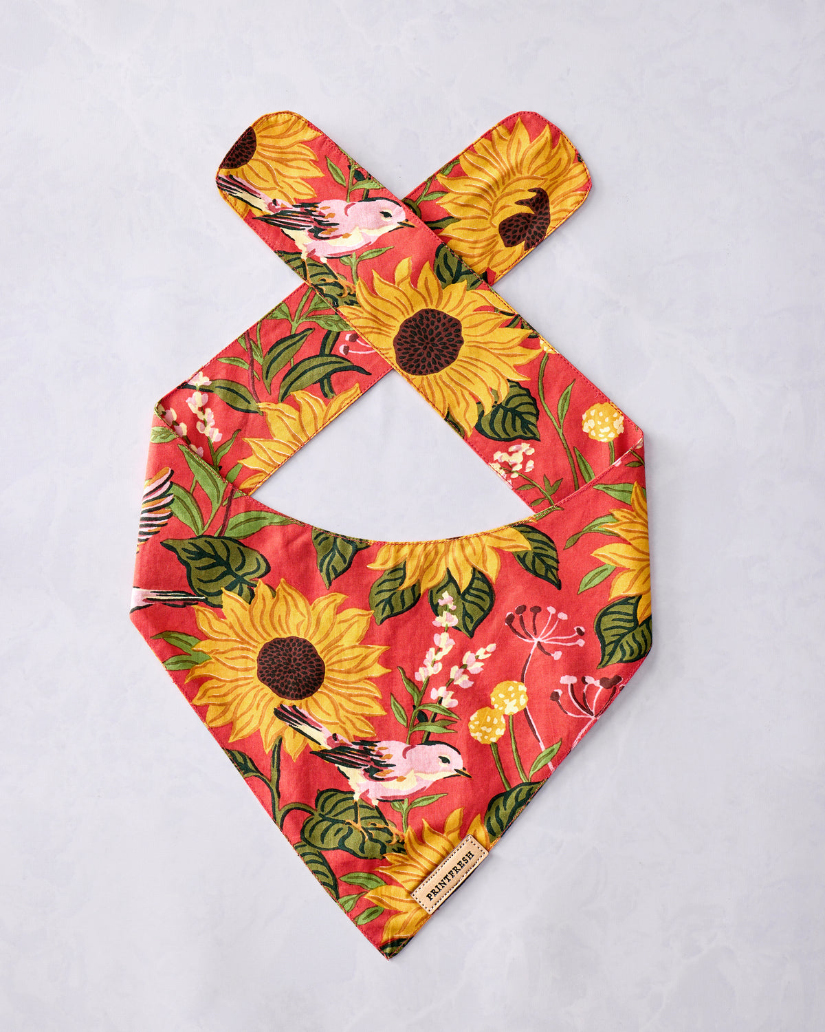 Orange pet bandana with crossed neck ties and a print of sunflowers, birds, leaves, and small flowers.