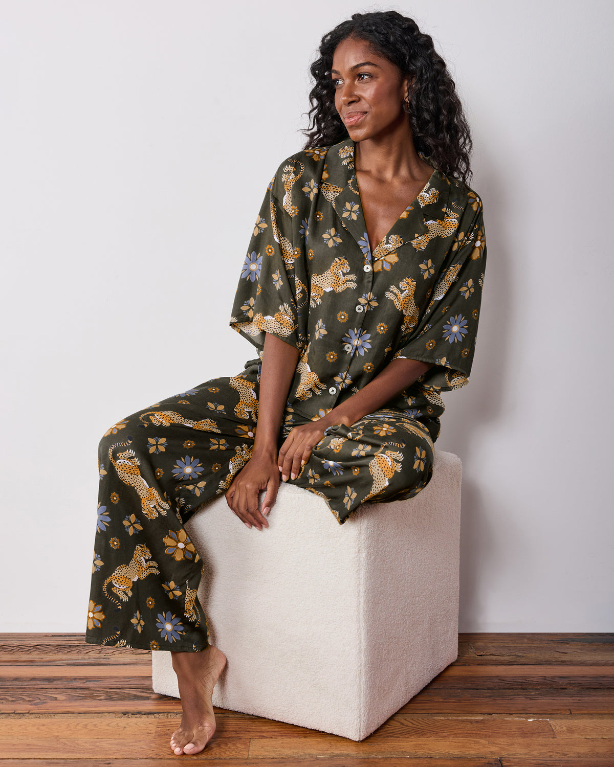 Seated front view of the olive Wildest Dreams pajama set, showing the short-sleeve button-front shirt and wide-leg pants with leopard and flower prints.