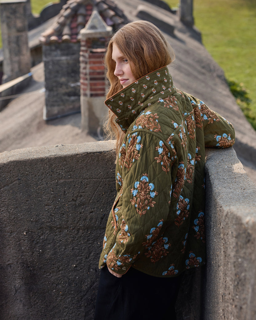 Back and side view of a quilted jacket in deep olive with a high collar and blue floral embroidery.