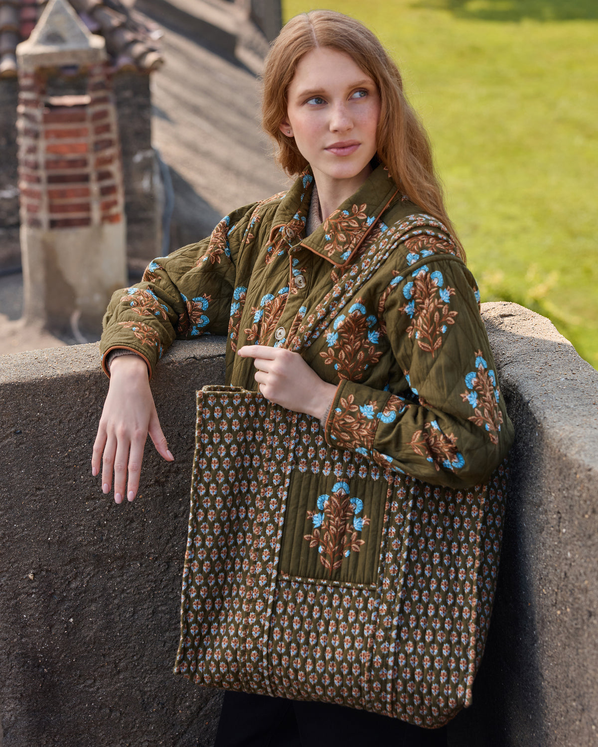 Front view of the deep olive quilted jacket and matching floral traveler tote, worn together.