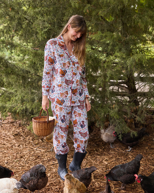 Person wearing a long-sleeved floral chicken-print pajama set with pants and boots among chickens outdoors.