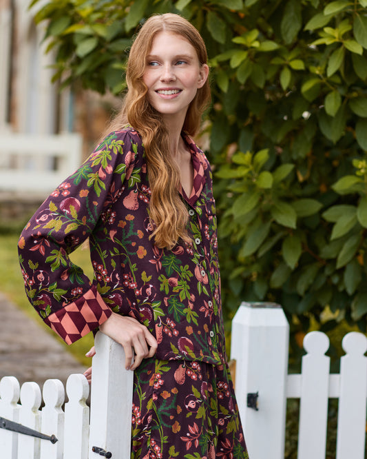 Close lifestyle view of the plum Pomme & Poire pajama top showing its button-front design, collar, chest pocket, fruit-and-leaf print, and contrasting geometric cuffs.