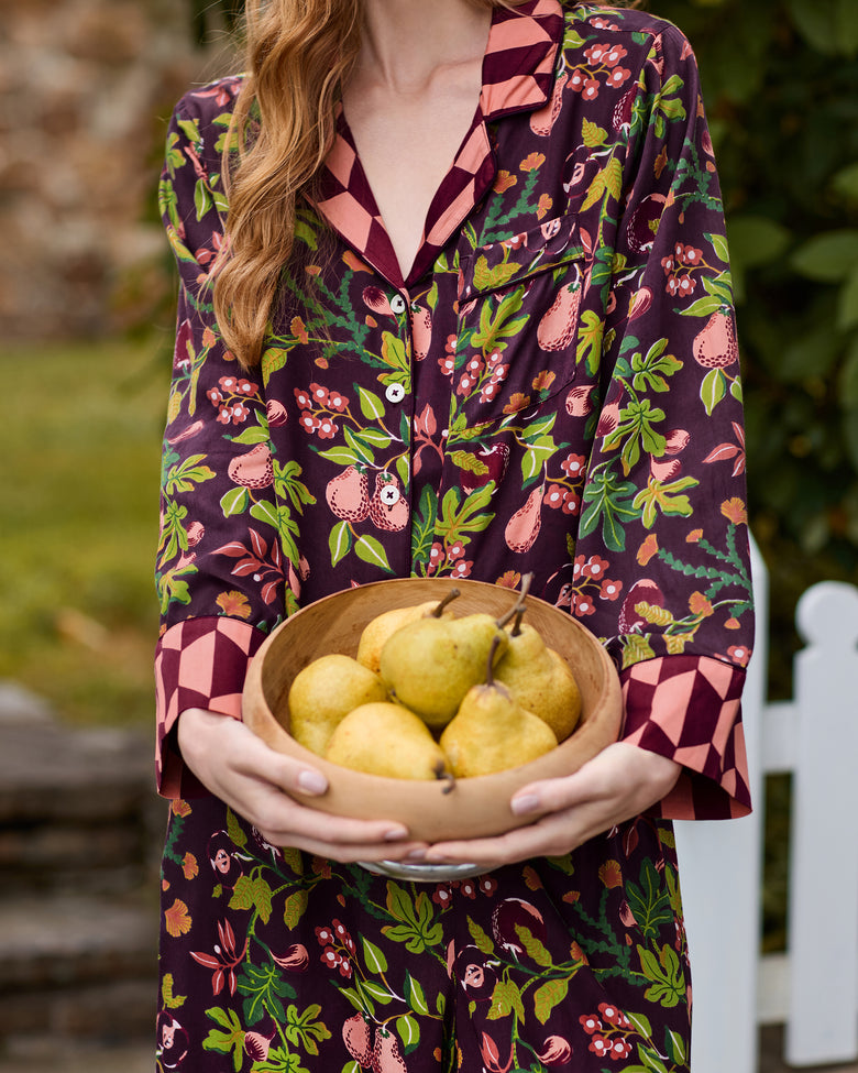 Close view of the plum Pomme & Poire pajama top and pants while a person holds a wooden bowl of pears; the fruit-and-leaf print and contrasting geometric cuffs are visible.