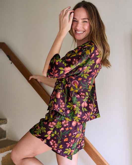 Person wearing the plum Pomme & Poire short pajama set with a long-sleeved button-front top and shorts, shown indoors beside a wooden stair rail; the set has a fruit-and-leaf print.