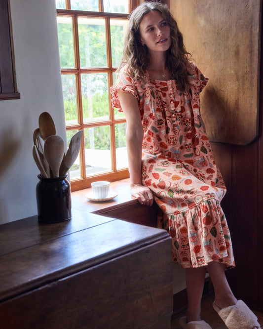 Front view of the Autumn Cafe nightgown worn seated on a window seat, showing its short sleeves, button front, and ruffled hem.
