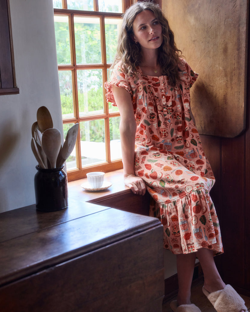 Front view of the Autumn Cafe nightgown worn seated on a window seat, showing its short sleeves, button front, and ruffled hem.