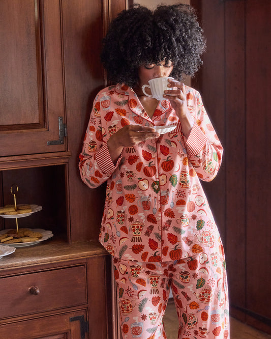Person wearing the pink Autumn Cafe long pajama set while drinking tea from a cup and holding a saucer; teapot, cup, fruit, leaf, and biscuit-jar print is visible.