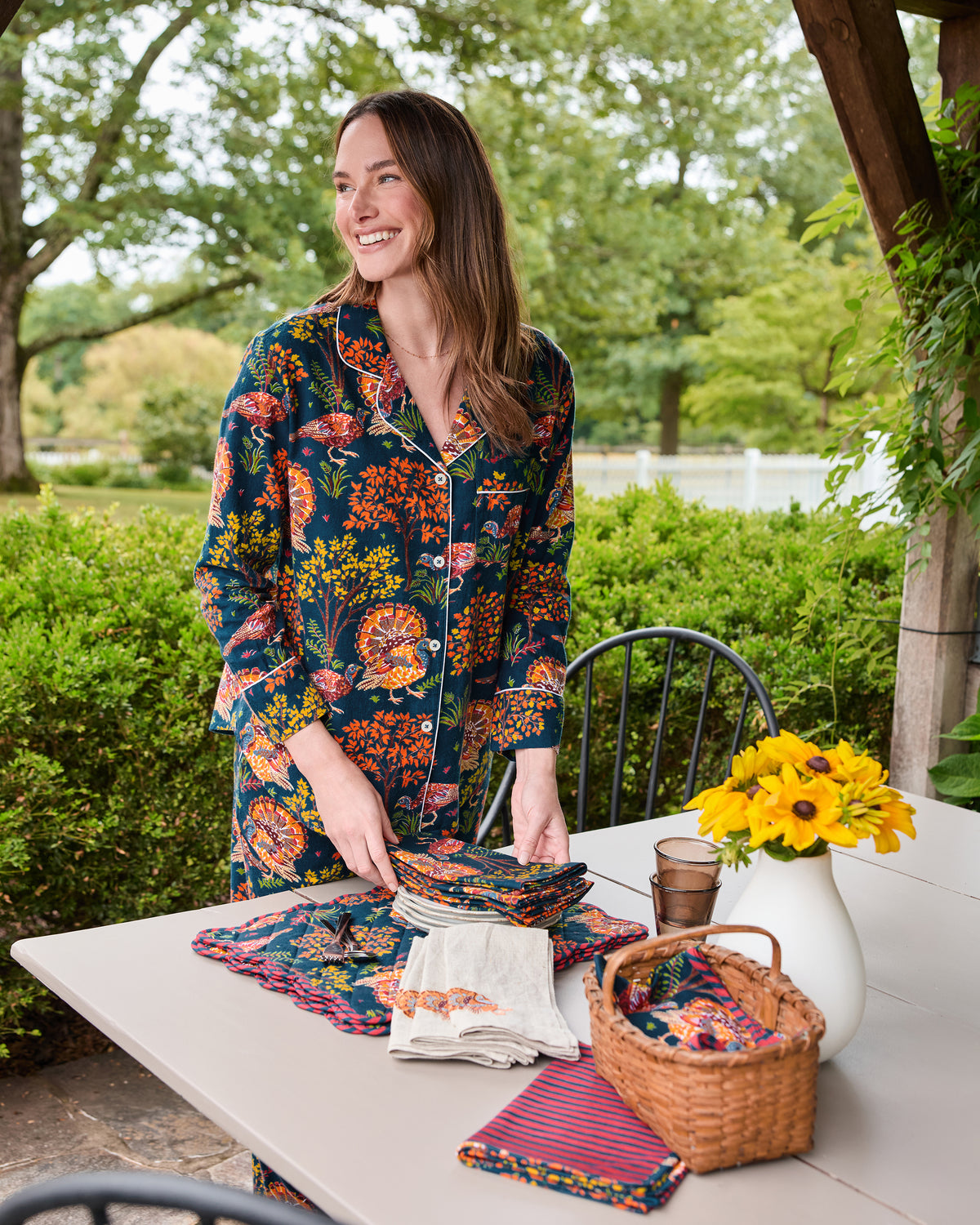 Person wearing the navy Turkey Trot long pajama set beside a table displaying coordinating folded textiles, napkins, a basket, and yellow flowers.