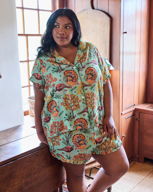 Person wearing a soft sage Turkey Trot short pajama set with turkey and autumn foliage print, ruffled sleeves, and ruffled shorts hem.